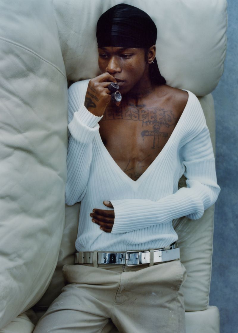 Portrait of a slender African-descent man with tattoos, wearing a black durag and low-cut ribbed white sweater, reclining on a white puffy couch.