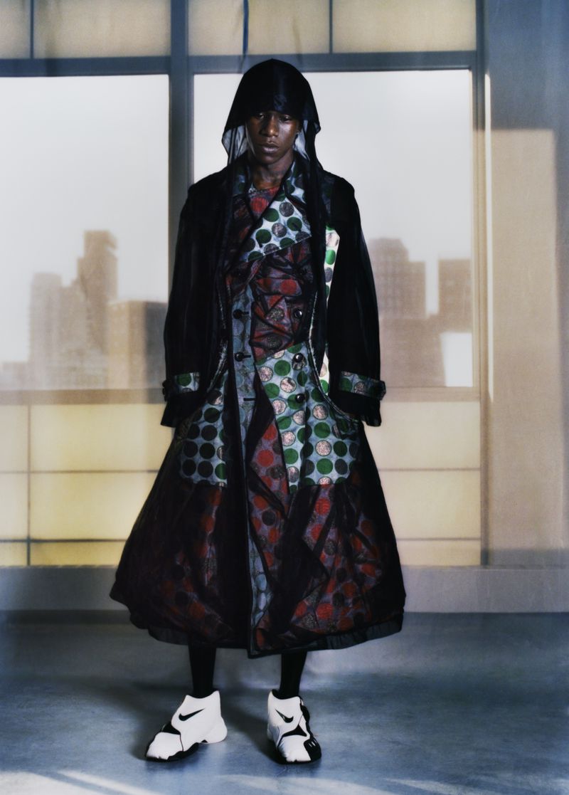 Full shot of a person in a hooded black coat and patterned inner coat, paired with athletic sneakers. Background of neutral window panes.