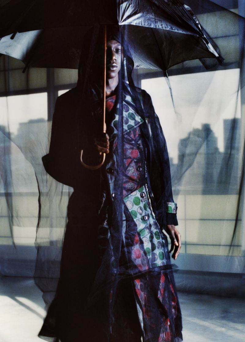 Fashionable adult in a semi-transparent coat and holding an umbrella stands against a city backdrop viewed through a window.
