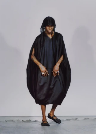 Model wearing a black hooded robe and dark shoes posing against a neutral grey studio backdrop