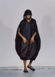 Model wearing a black hooded robe and dark shoes posing against a neutral grey studio backdrop