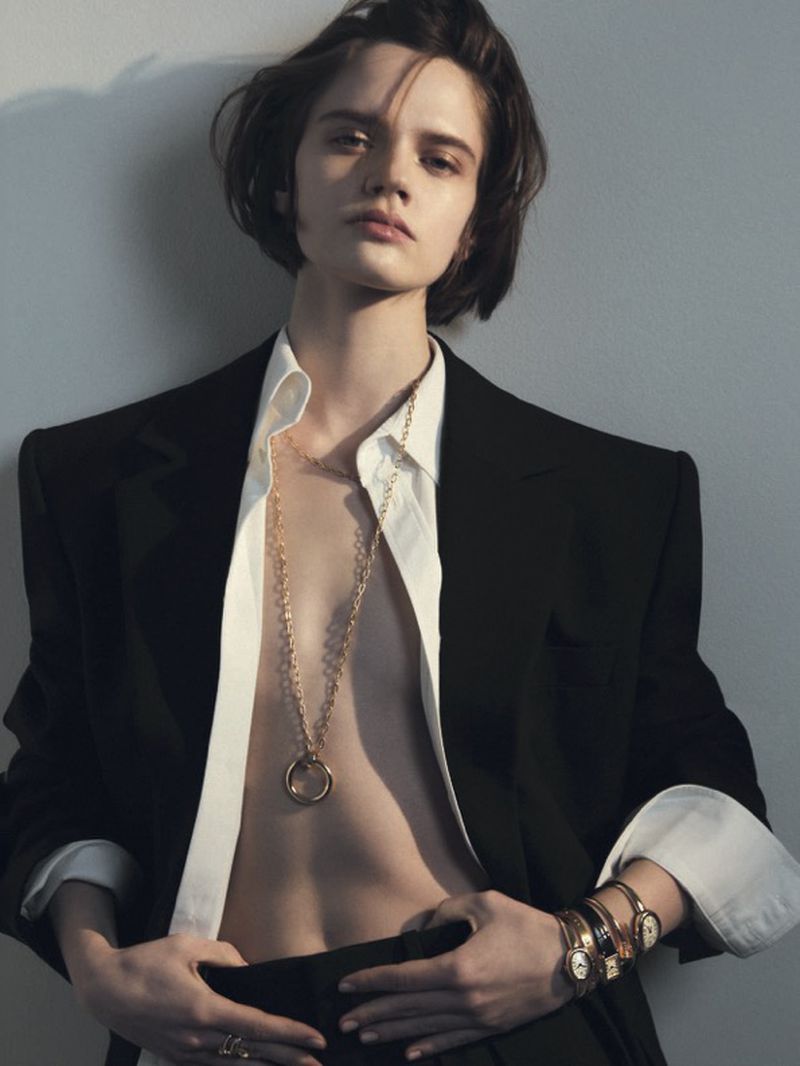 A young woman poses in a white shirt and black blazer, accentuated by a gold necklace and bracelets, set against a neutral background.
