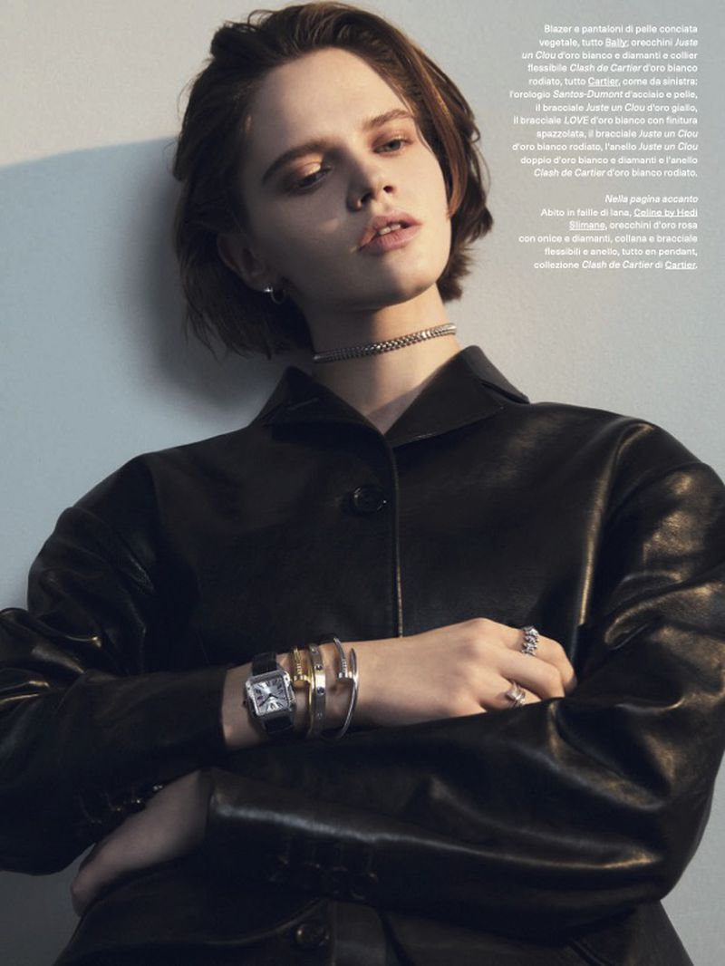 Fashion editorial portrait of a woman in a black leather jacket with jewelry, posing indoors with a neutral wall backdrop.