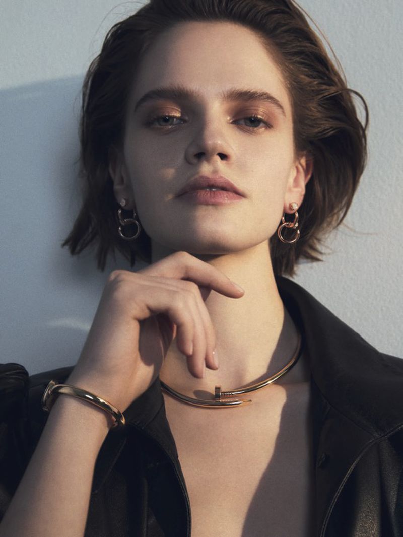 Portrait of a woman with short brown hair wearing gold jewelry and a black leather jacket in a brightly lit, neutral setting.
