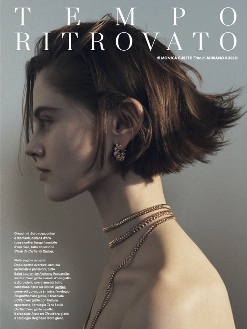 A woman is featured in a close-up studio shot. She's adorned with gold jewelry and has short brown hair, captured in soft, diffused lighting.