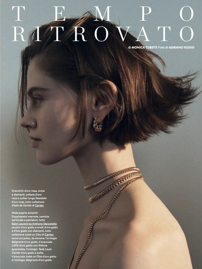 Profile portrait of a short-haired model wearing gold chain necklaces and hoop earrings against a plain background