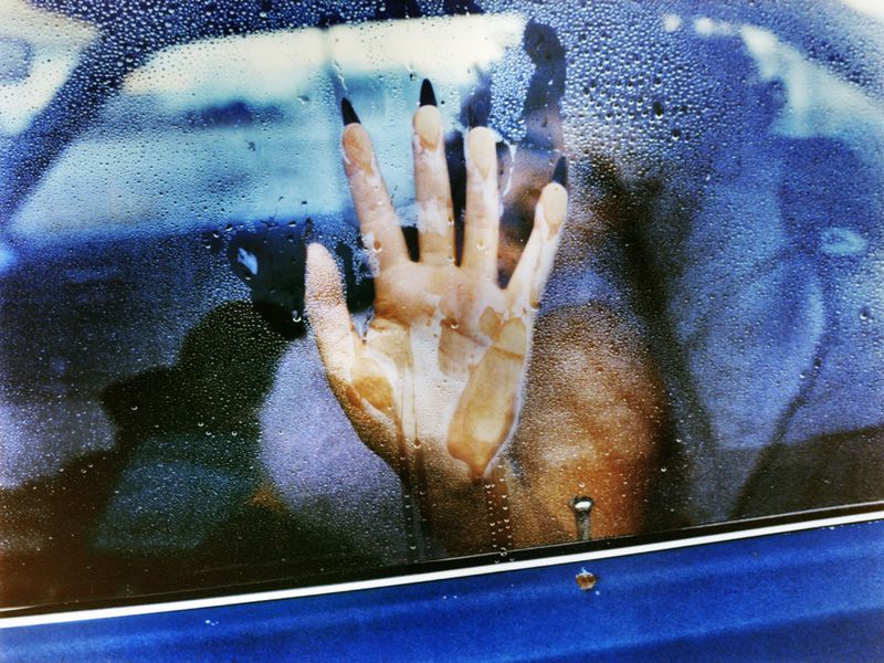 Close-up view through a wet car window showing a woman's hand with long black fingernails pressed against the glass, creating water streaks.