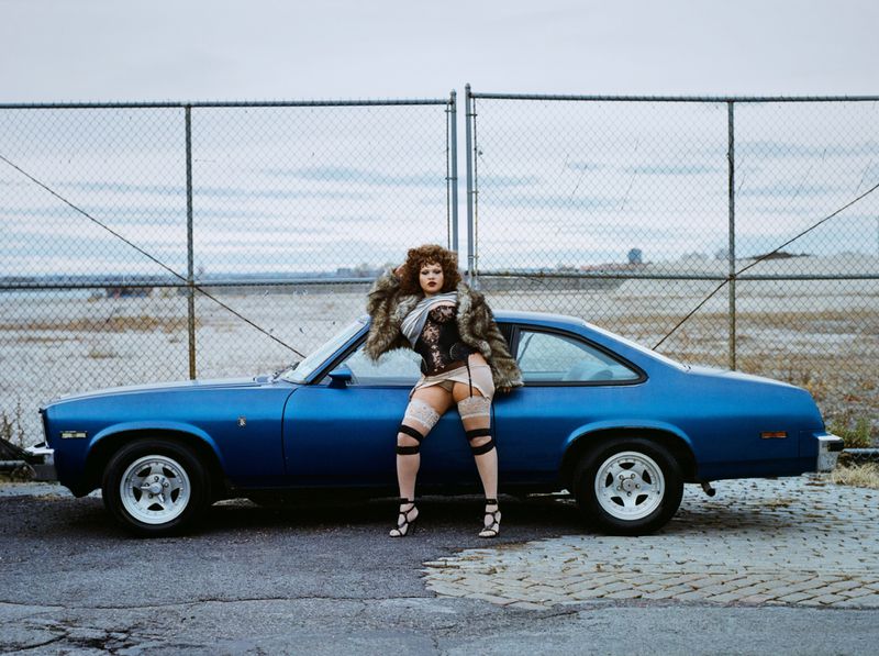 A vintage blue car serves as a striking backdrop for a woman in a fur coat, corset, and gartered thigh-highs, set against a chain-link fence.