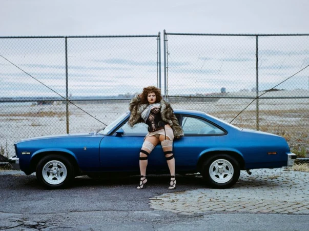 A vintage blue car serves as a striking backdrop for a woman in a fur coat, corset, and gartered thigh-highs, set against a chain-link fence.