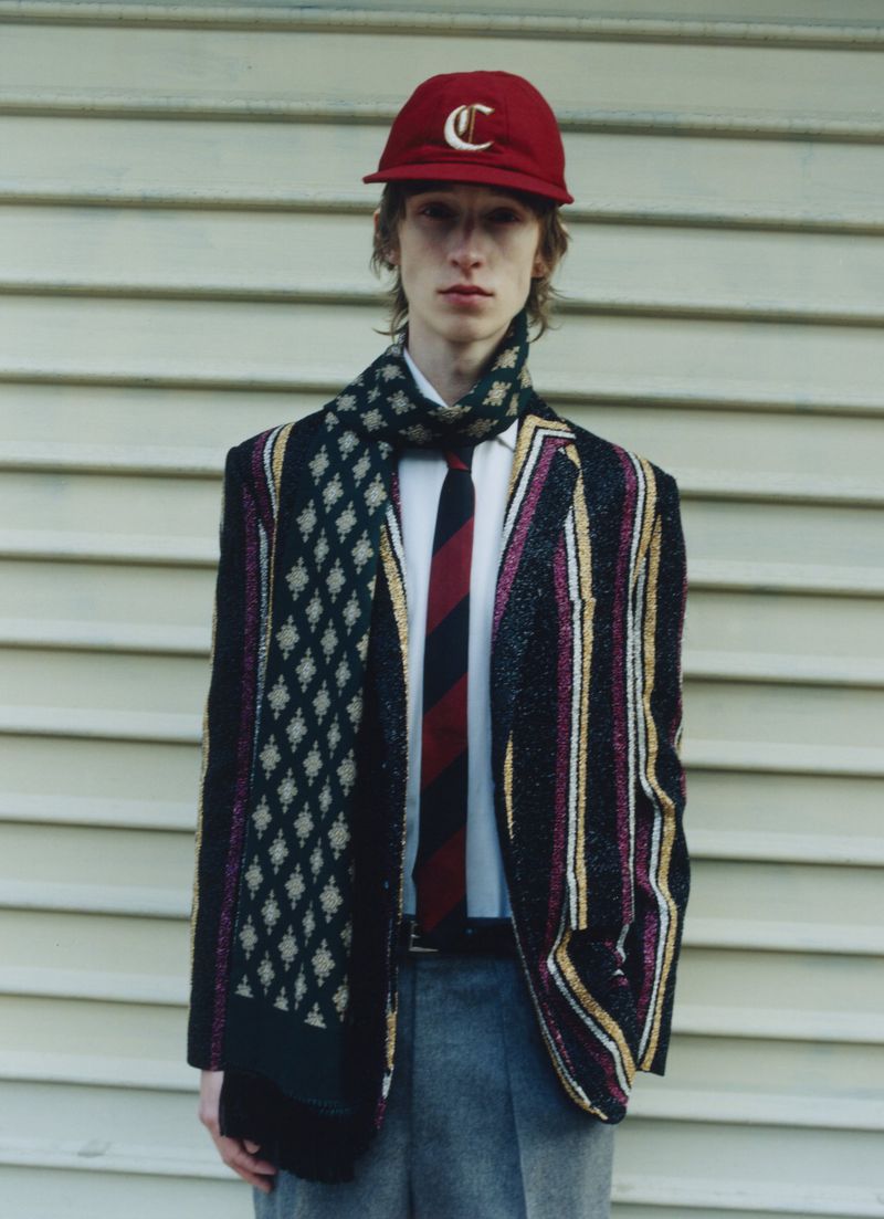 Young adult man wearing a red baseball cap, scarf, blazer, dress shirt, and tie, posing in front of a horizontal siding wall.