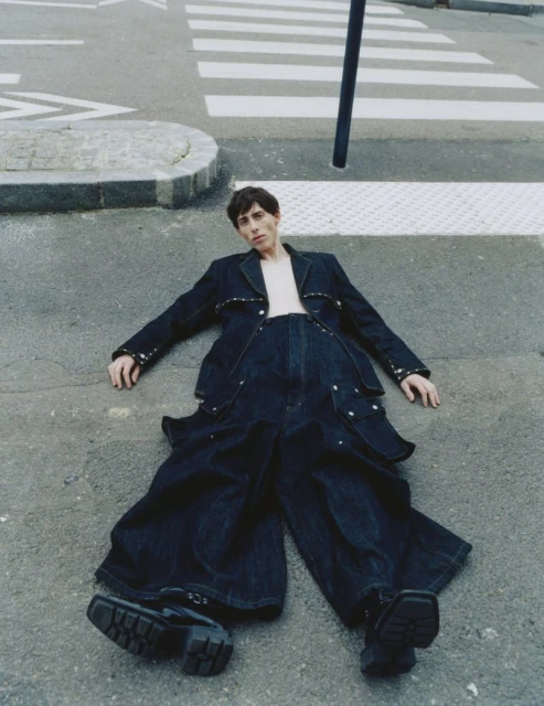 Fashion shot of a young adult lying on grey asphalt wearing an oversized dark denim outfit and chunky black shoes, with a crosswalk in the background.