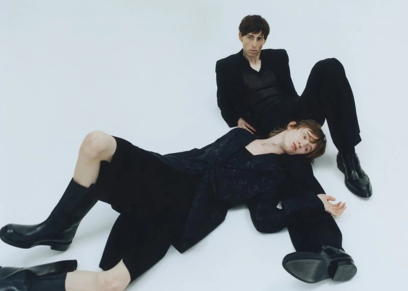 Two fashion models in dark apparel, one lying down and one sitting, pose against a white backdrop in a studio setting.