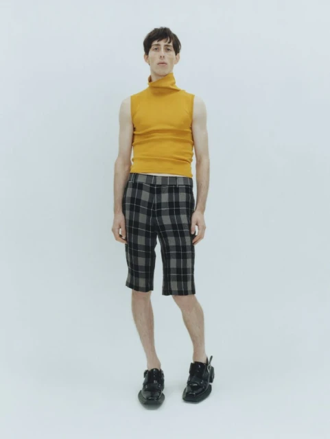 Full-length shot of an adult male model in a sleeveless gold turtleneck, plaid shorts, and black shoes against a white backdrop.