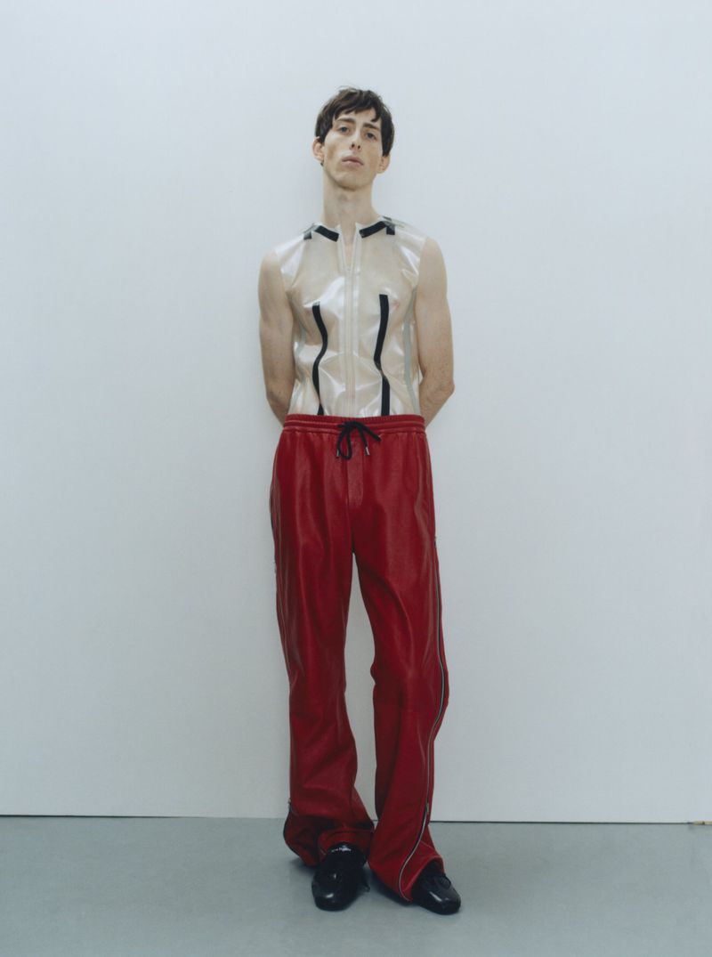A slender adult male fashion model in a studio setting wears a transparent top and red sweatpants, standing with arms behind his back.