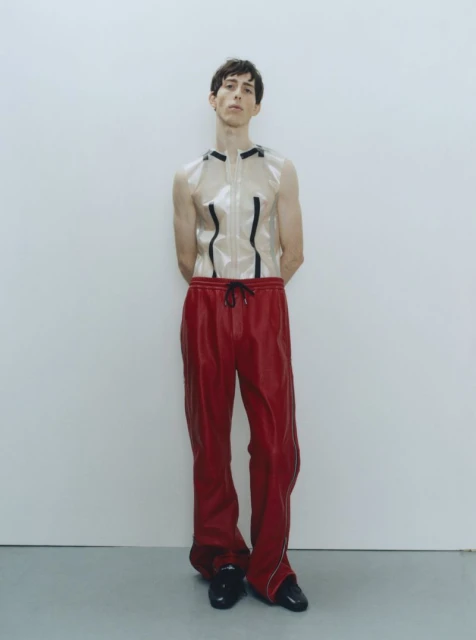 A slender adult male fashion model in a studio setting wears a transparent top and red sweatpants, standing with arms behind his back.