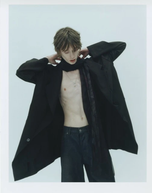 A slender person with tousled hair poses in a black coat, black jeans, and a scarf against a bright white photo studio backdrop.