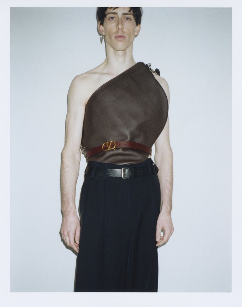 A male model wearing a sculptural brown asymmetrical top and dark trousers against a white seamless studio backdrop.