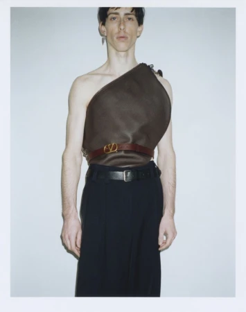 A male model wearing a sculptural brown asymmetrical top and dark trousers against a white seamless studio backdrop.