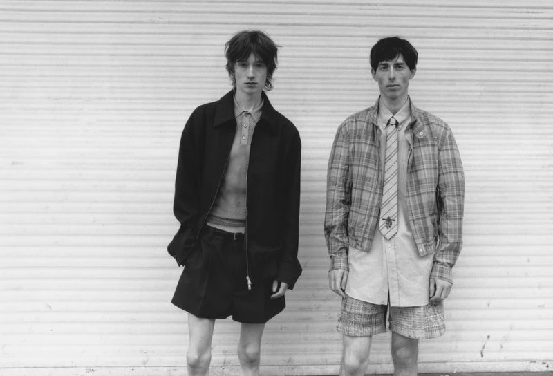 Two young men pose outdoors against a white shutter door, showcasing fashion with jackets, shirts, ties, and shorts in a monochrome aesthetic.