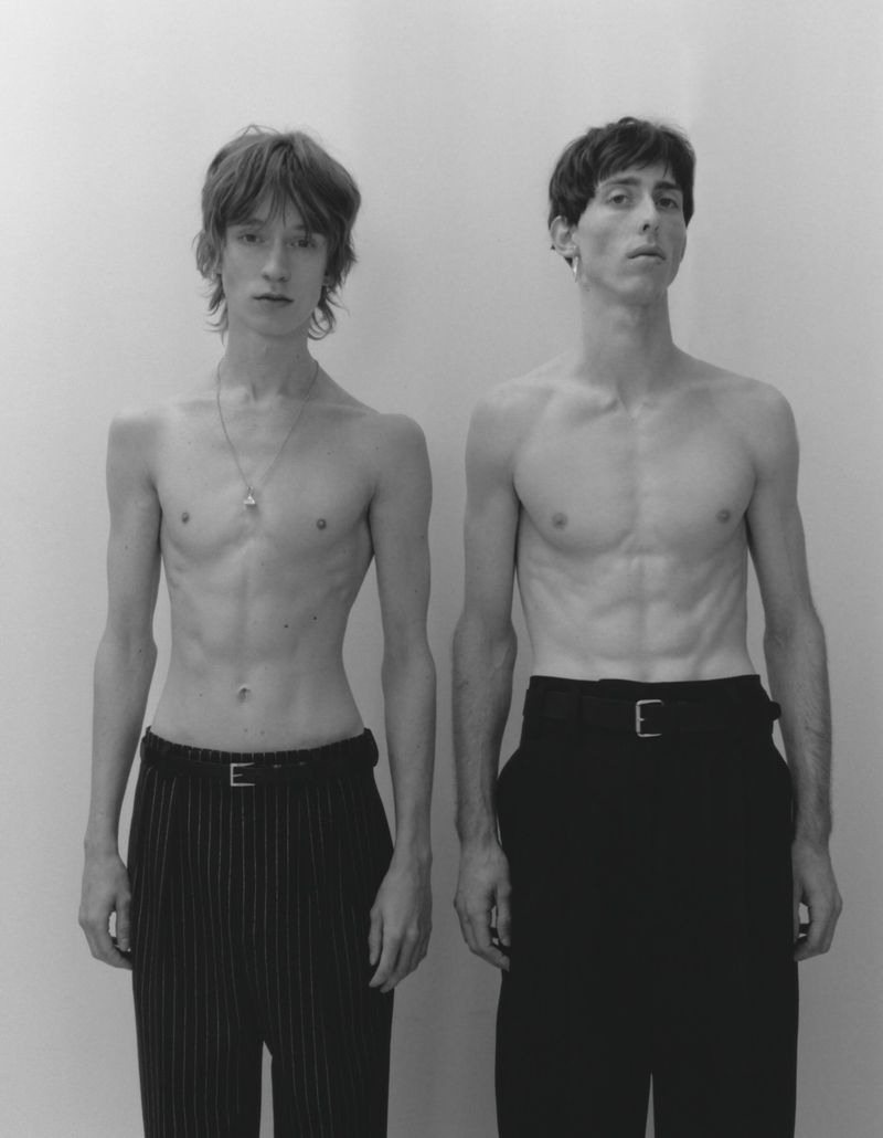 Two shirtless male models standing side by side wearing dark trousers against a plain light studio background.