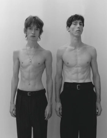 Two shirtless male models standing side by side wearing dark trousers against a plain light studio background.