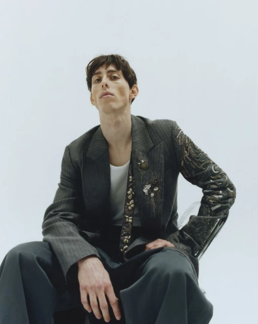 A person in a grey pinstripe suit jacket with embellishments poses in a studio, seated on a transparent chair against a white backdrop.