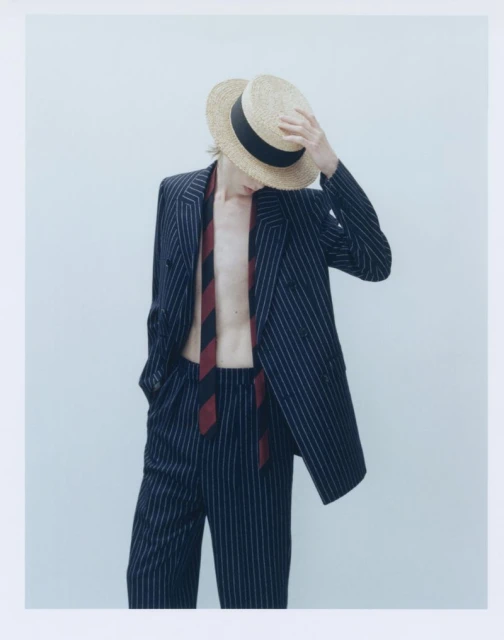A person wearing a pinstripe suit and boater hat is covering their face with a hand in a studio setting with a light backdrop.