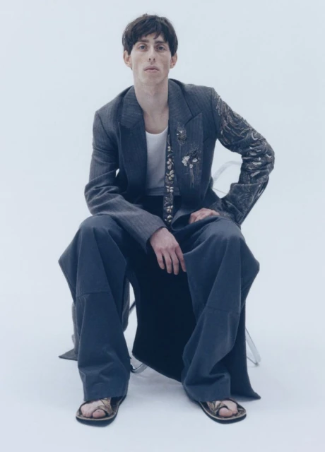 Male fashion model in an oversized grey suit and sandals sits on a clear chair in a bright photo studio, posing in a direct gaze.