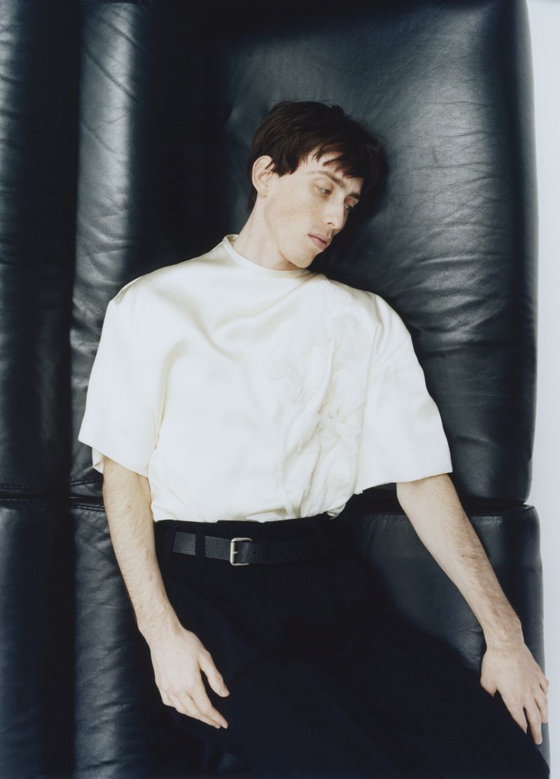 A young male model lying against a dark leather couch wearing a cream t-shirt and black trousers with a belt.