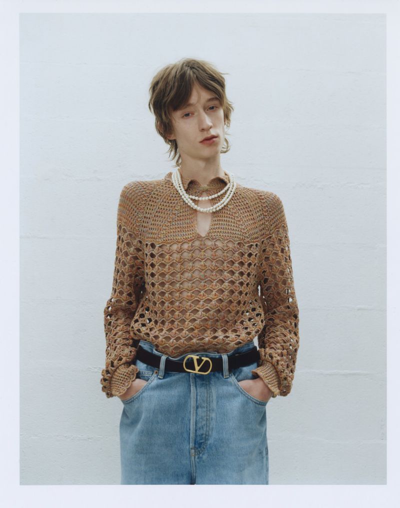 Adult male in a light brown crochet sweater, pearl necklace, light blue jeans and a black belt poses in front of a white wall.