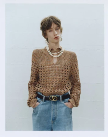 Adult male in a light brown crochet sweater, pearl necklace, light blue jeans and a black belt poses in front of a white wall.