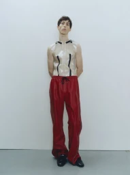 Male model in a translucent sleeveless top and red trousers standing against a plain light studio background.