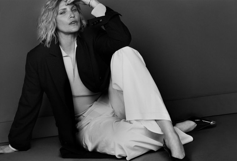Monochrome portrait of a blonde woman in an oversized blazer and white pants sitting indoors with a hand to her forehead.