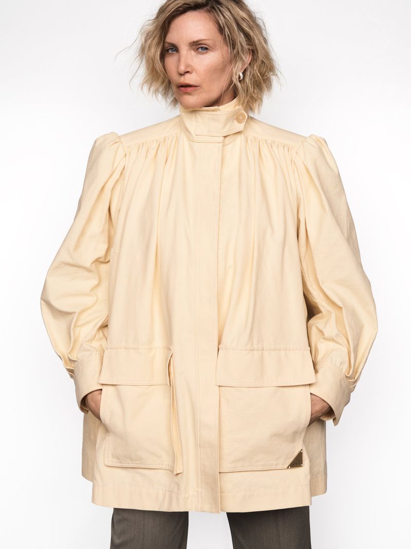 Woman in a light yellow coat with large sleeves posing in front of a white background in a fashion studio shot.