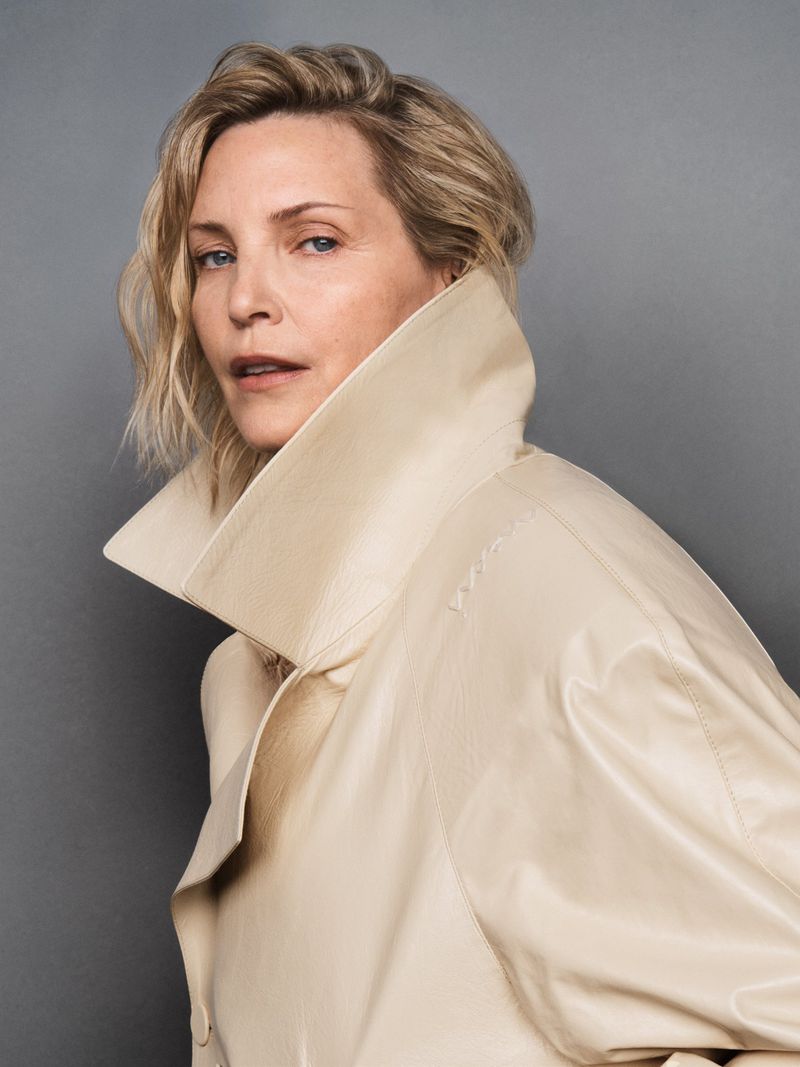 Blonde woman in a cream-colored leather coat, portrait shot against a grey backdrop, showcasing high fashion and studio photography.