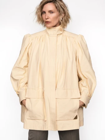 Woman with short blonde hair wearing a light yellow high collar jacket against a white background