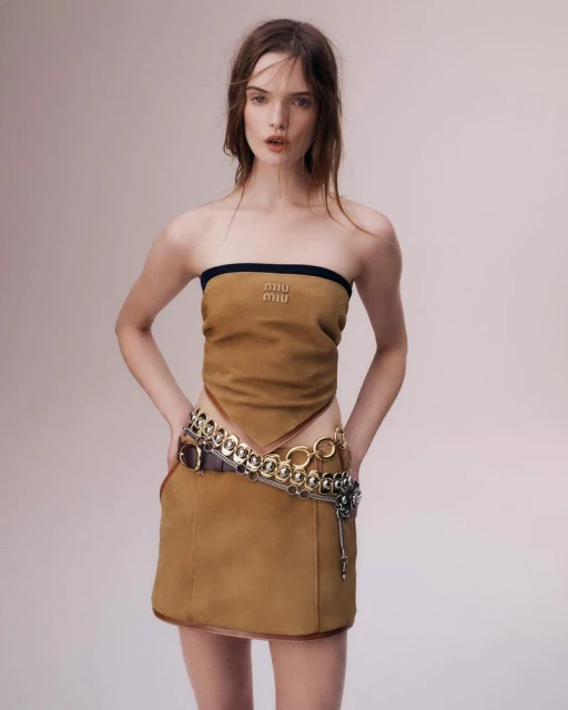 A young adult female fashion model poses in a tan Miu Miu tube top and tan skirt with a silver chain belt, standing in a photo studio.