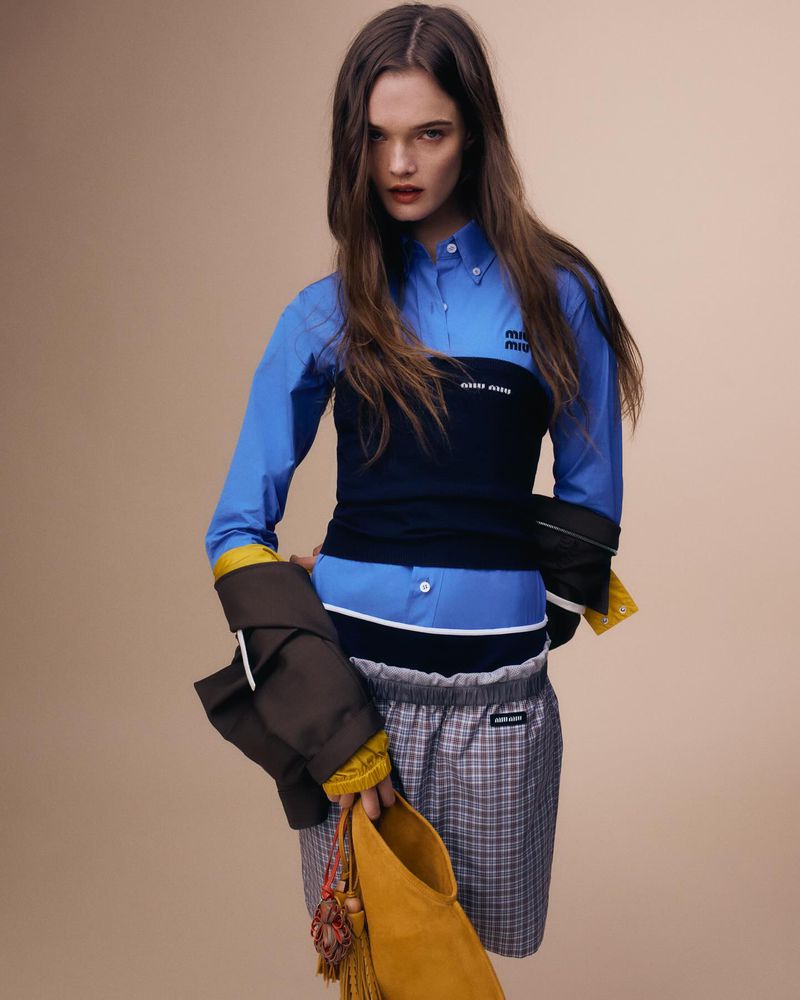 Fashion editorial shot of a brunette woman in a blue shirt and plaid shorts holding a brown jacket and yellow purse in a studio.