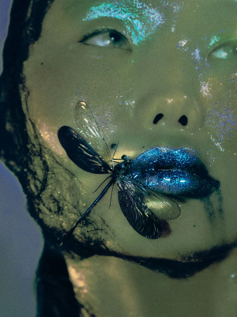 Close-up portrait of person with glitter makeup, dragonfly on lips. Artistic, glamourous beauty shot with dramatic lighting and bold styling.