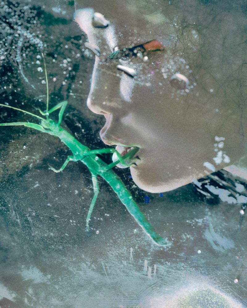 Close-up surreal portrait of an adult female with a bright green stick insect on her lip in front of a blurred neutral background.