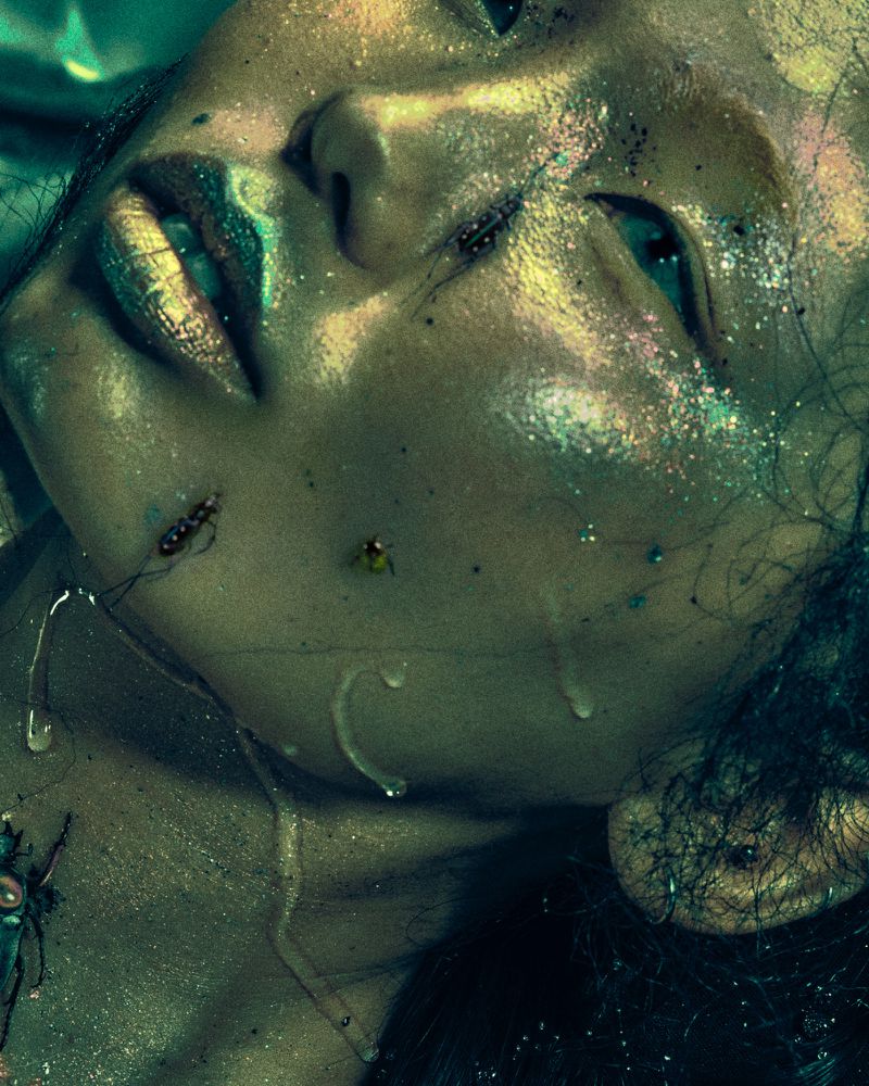 Artistic beauty shot of a fair-skinned woman with glitter makeup and prop insects against a dark background with water droplets.