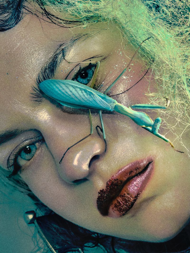 A close-up portrait shot of a fair-skinned adult female model with a striking green mantis insect on her face, beauty lighting, and fashion.