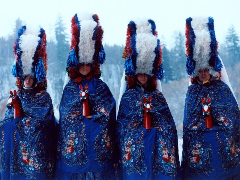 Four people in blue patterned robes and tall, colorful hats stand in a snowy outdoor setting with trees in the background.