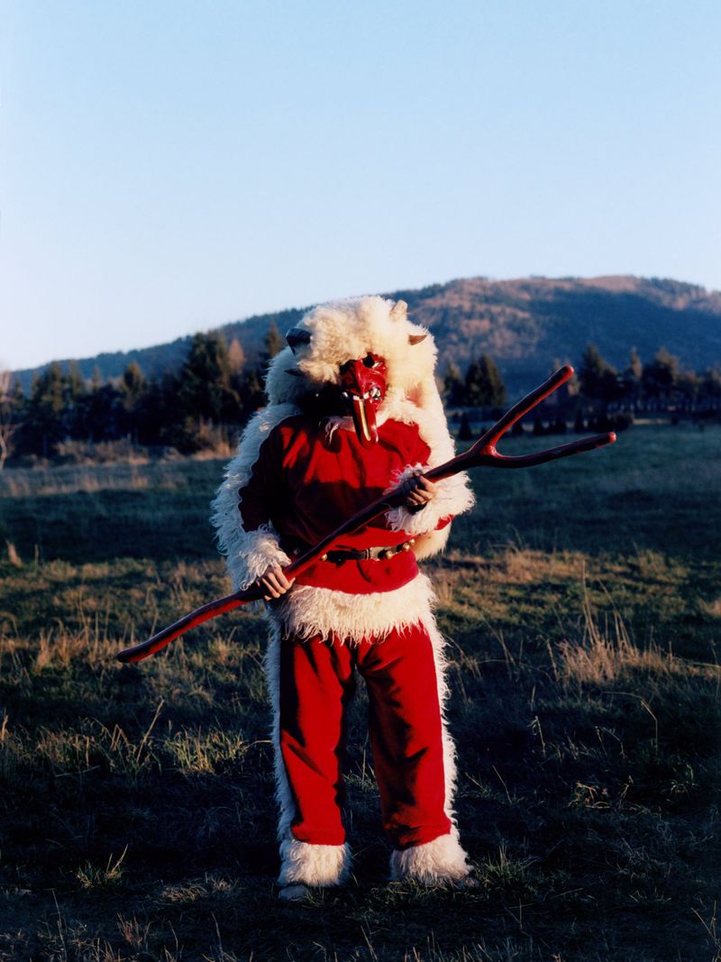 Person in red and white Krampus costume with mask holding a wooden staff stands in a field with hills and sky in the background.