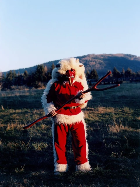 Person in red and white Krampus costume with mask holding a wooden staff stands in a field with hills and sky in the background.
