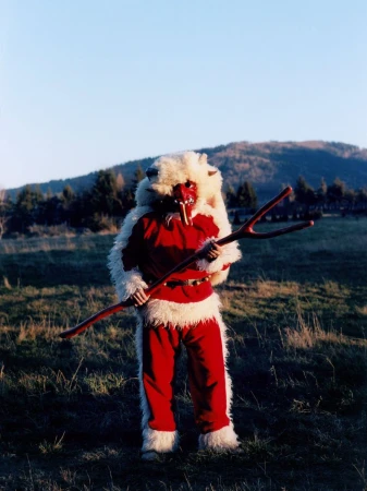 Person in red and white Krampus costume with mask holding a wooden staff stands in a field with hills and sky in the background.