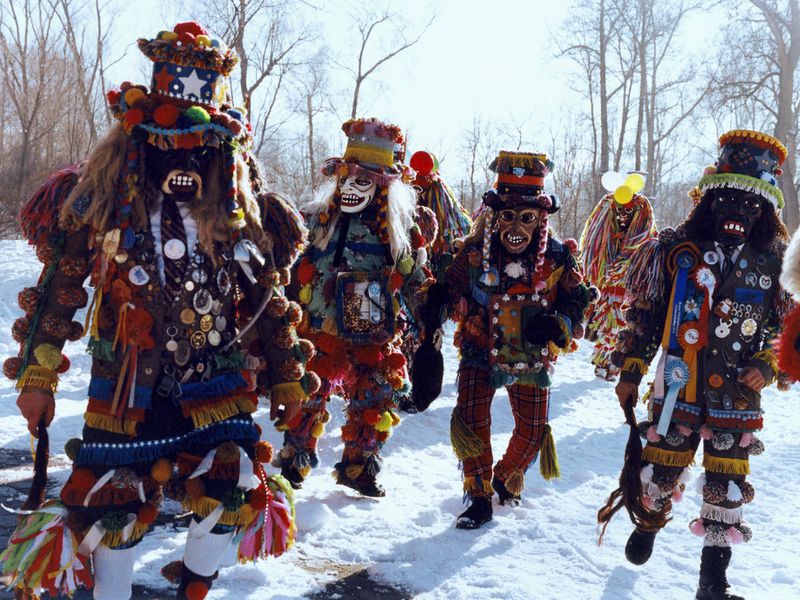 Mummers in elaborate costumes parade through snow in daylight. Street scene with masked and costumed figures and trees in background.