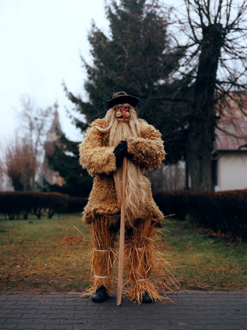 A person in a traditional costume with straw and fur stands on a stone path in a village, with a church tower visible in the background.
