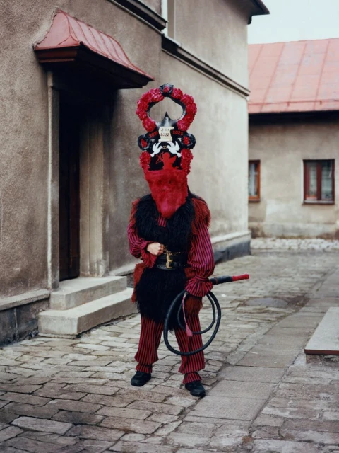 Person in a Perchtenlauf costume with a whip stands outdoors against buildings in a cobblestone courtyard on an overcast day.