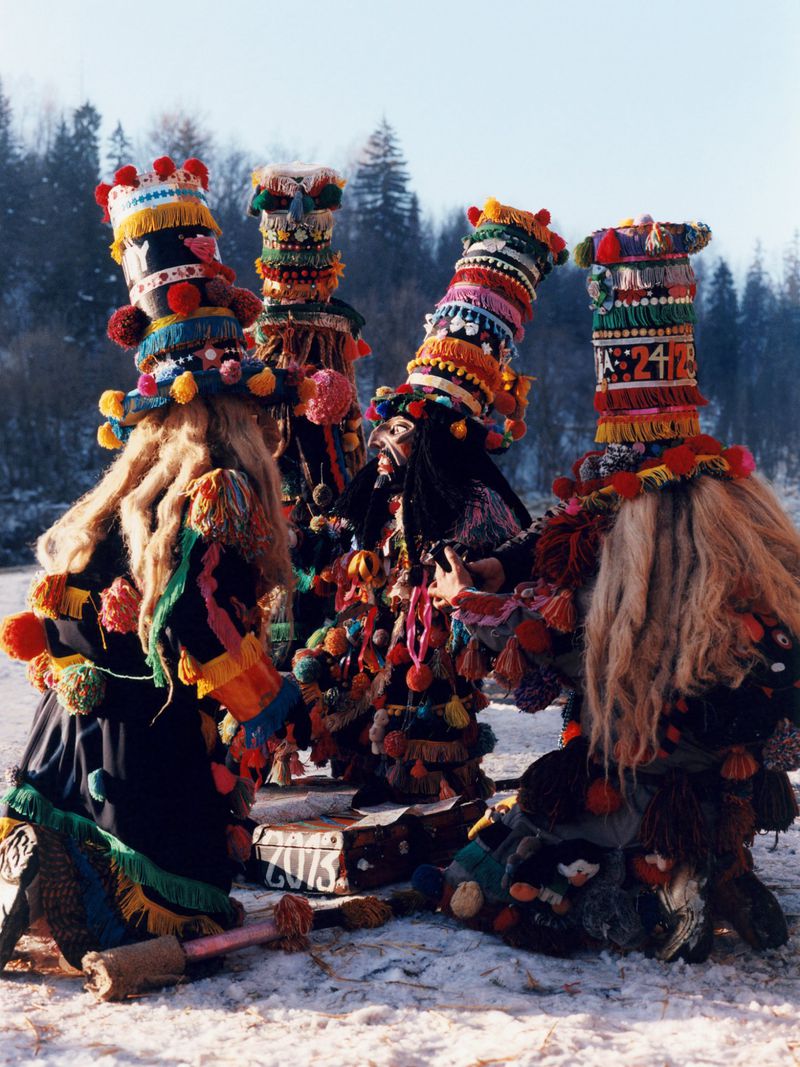 Five people in traditional carnival costumes with tall hats, tassels, and long fur, standing in a snowy field with trees in the background.